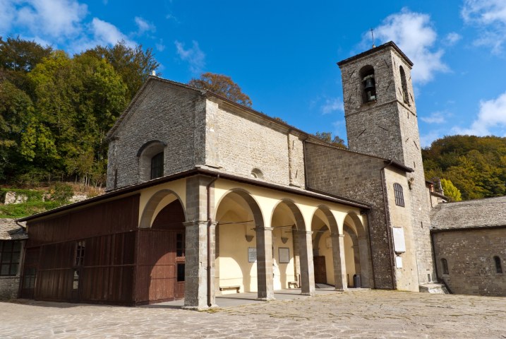 Chiesa in pietra con portico ad archi e campanile, circondata da boschi autunnali sotto un cielo azzurro